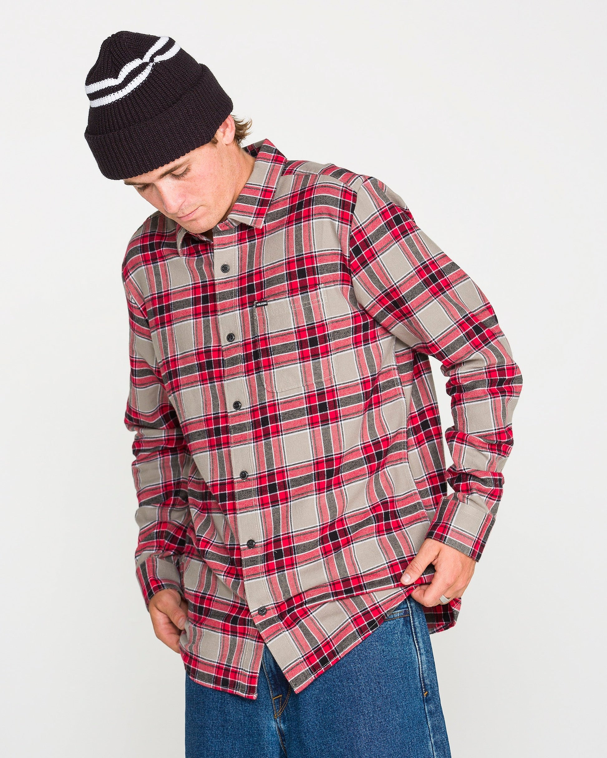 A person wearing the Volcom Men's Caden Plaid Long Sleeve Shirt in red and gray with classic fit, paired with blue jeans and a black knit beanie, looks down while adjusting the shirt hem against a plain white background.