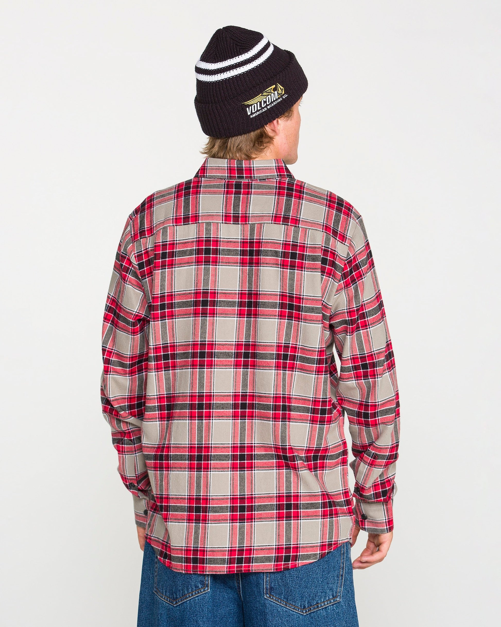 A person wearing a black beanie, blue jeans, and the Volcom Men's Caden Plaid Long Sleeve Shirt in a classic fit red and beige cotton plaid is standing with their back to the camera against a plain white background.