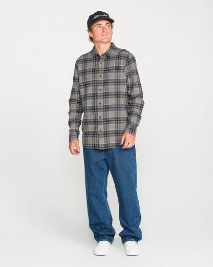 A man wearing a black cap, blue jeans, and white sneakers stands against a plain white background, looking slightly to his left. He is dressed in the Volcom Men's Caden Plaid Long Sleeve Shirt in a classic fit.