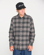A man models the Volcom Men's Caden Plaid Long Sleeve Shirt in classic fit gray and dark blue, paired with a black cap and blue jeans, standing against a plain white background and looking slightly to the side.