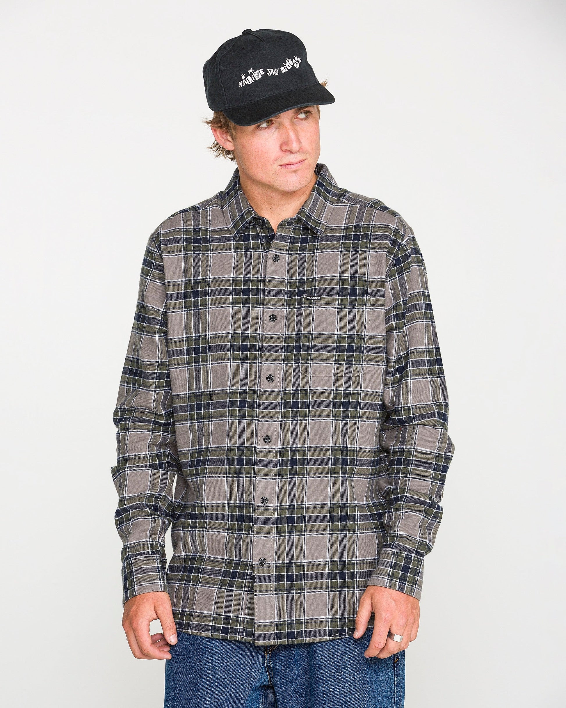 A man models the Volcom Men's Caden Plaid Long Sleeve Shirt in classic fit gray and dark blue, paired with a black cap and blue jeans, standing against a plain white background and looking slightly to the side.