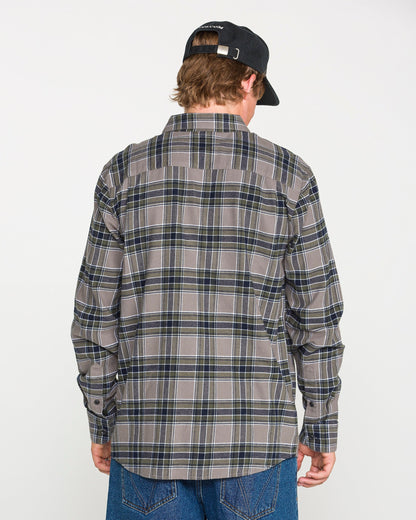 A person is standing with their back to the camera against a plain white background, wearing a black cap, blue jeans, and the Volcom Men's Caden Plaid Long Sleeve Shirt in a classic fit gray and blue plaid.
