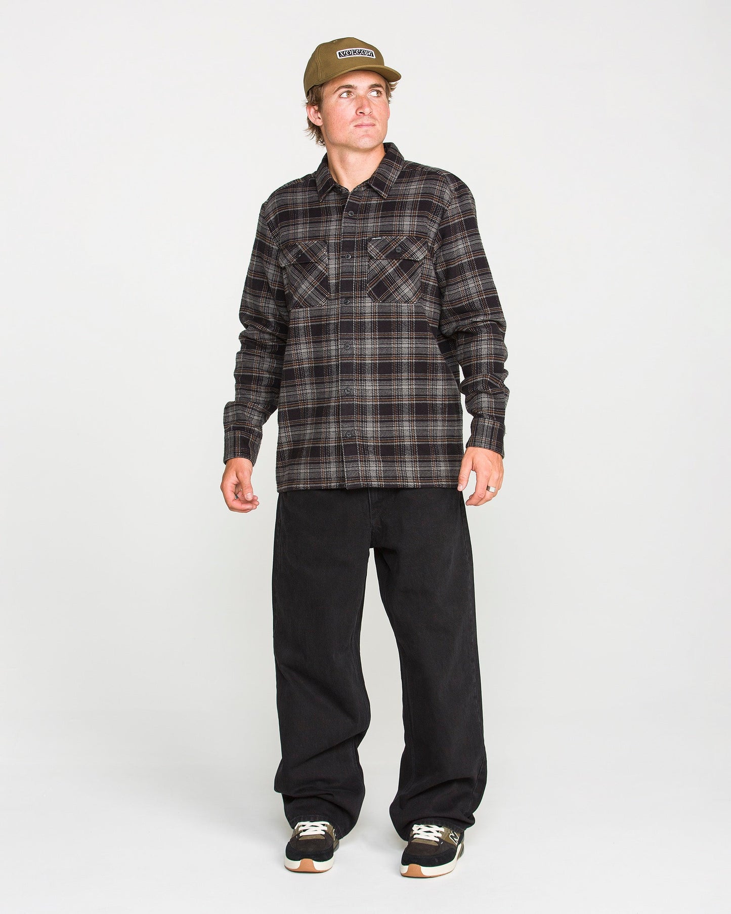 A person wearing the Volcom Men's Johnstone Long Sleeve Shirt in brown plaid, paired with black pants, black sneakers, and a tan cap stands against a plain white background, looking slightly to their left. The shirt's brushed flannel fabric adds texture and warmth to their casual look.