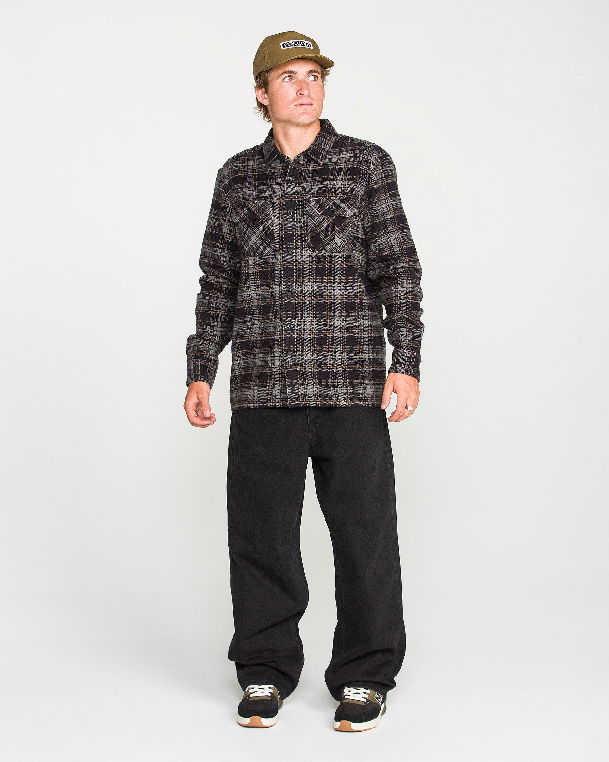 A person wearing the Volcom Men's Johnstone Long Sleeve Shirt in brown plaid, paired with black pants, black sneakers, and a tan cap stands against a plain white background, looking slightly to their left. The shirt's brushed flannel fabric adds texture and warmth to their casual look.