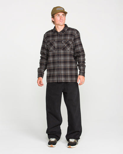 A person wearing the Volcom Men's Johnstone Long Sleeve Shirt in brown plaid, paired with black pants, black sneakers, and a tan cap stands against a plain white background, looking slightly to their left. The shirt's brushed flannel fabric adds texture and warmth to their casual look.