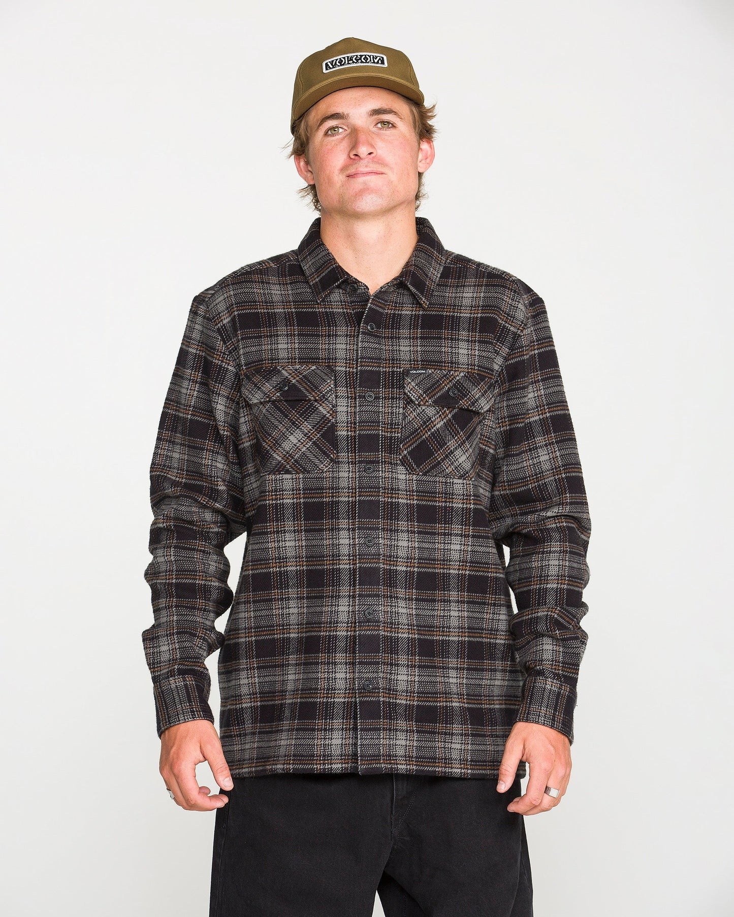 A person wearing a beige cap, black pants, and the Volcom Men's Johnstone Long Sleeve Shirt — a black-and-brown plaid heavyweight flannel with chest pockets — stands facing forward against a plain white background.