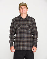 A person wearing a beige cap, black pants, and the Volcom Men's Johnstone Long Sleeve Shirt — a black-and-brown plaid heavyweight flannel with chest pockets — stands facing forward against a plain white background.