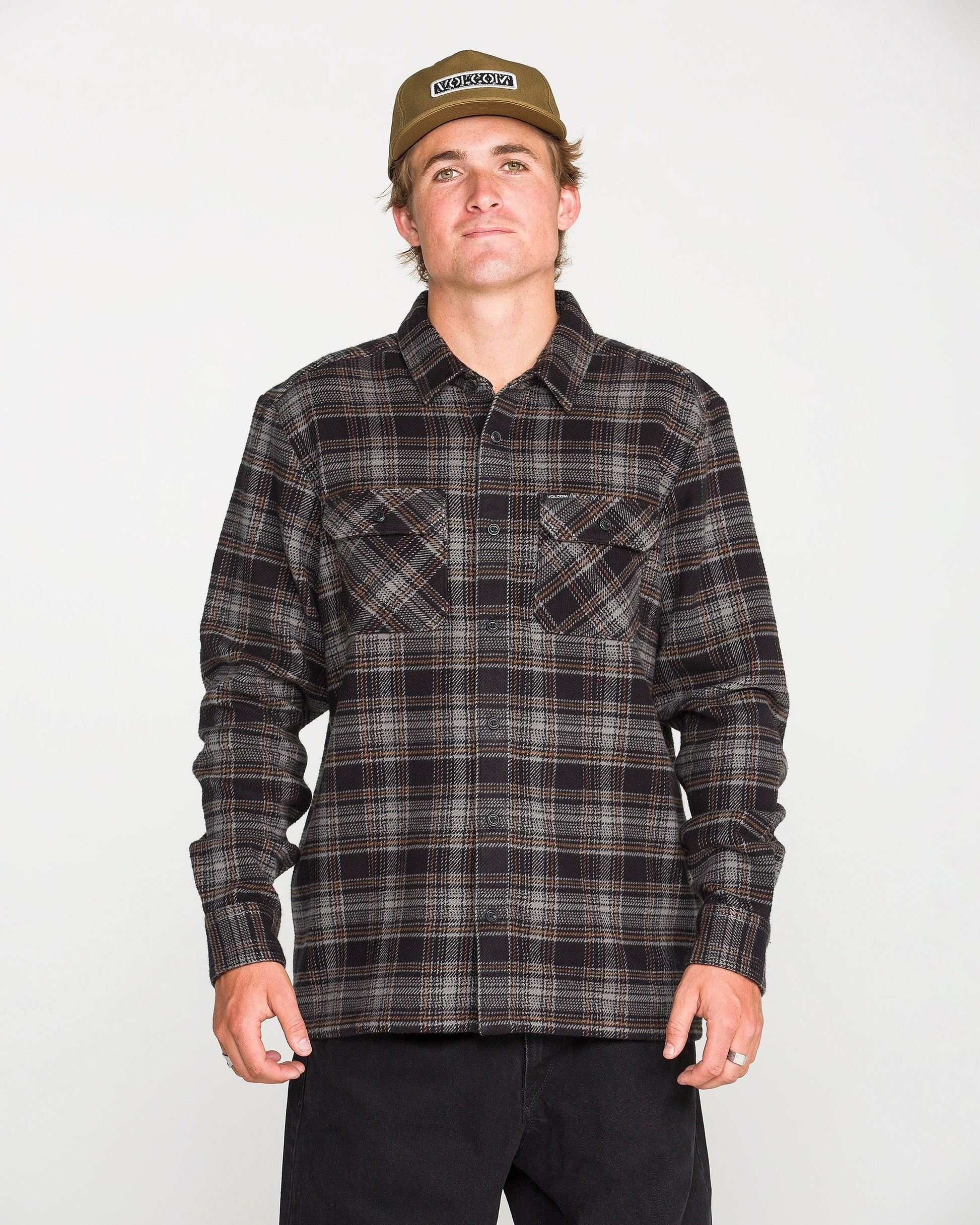 A person wearing a beige cap, black pants, and the Volcom Men's Johnstone Long Sleeve Shirt — a black-and-brown plaid heavyweight flannel with chest pockets — stands facing forward against a plain white background.