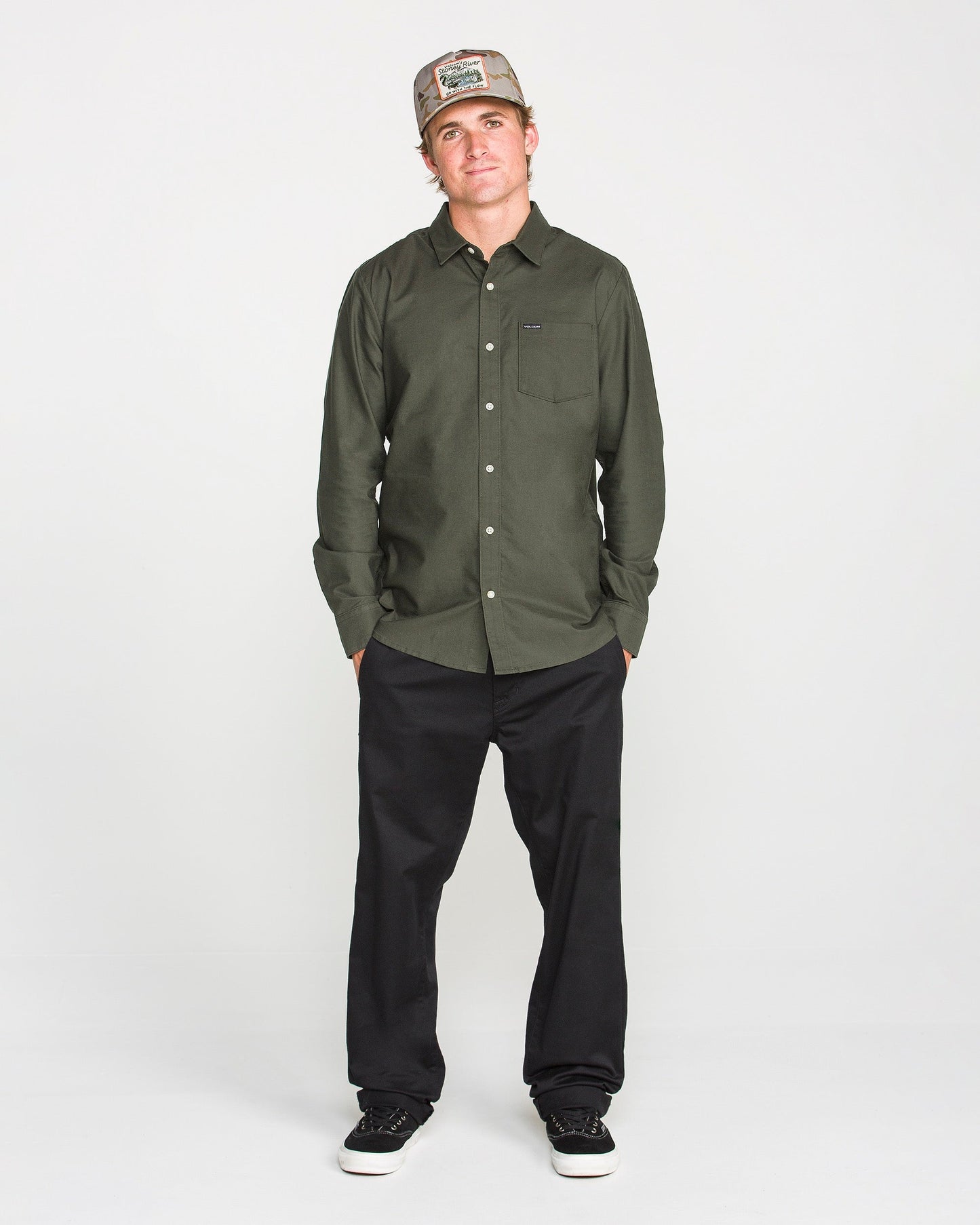 A man in a Volcom Men's Veeco Oxford Long Sleeve Shirt in Climbing Ivy green, classic fit black pants, and black sneakers stands with his hands in his pockets, wearing a tan baseball cap against a plain white background.