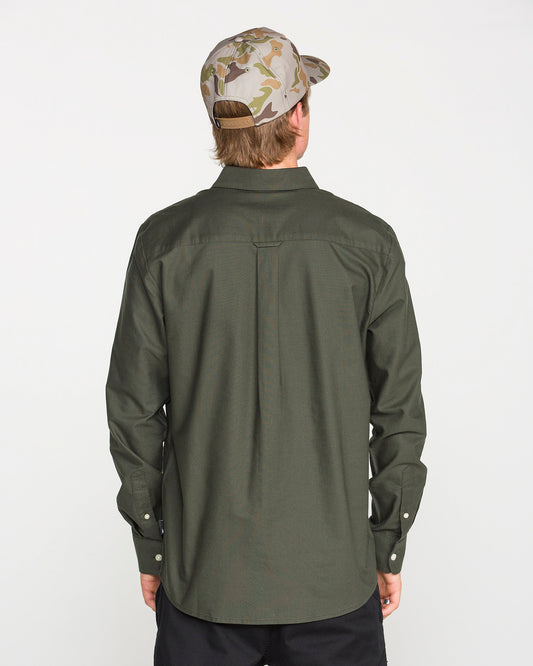 A person wearing a Climbing Ivy green Men's Veeco Oxford Long Sleeve Shirt in a classic fit by Volcom and a camouflage baseball cap stands facing away, displaying the back of their outfit against a plain white background.