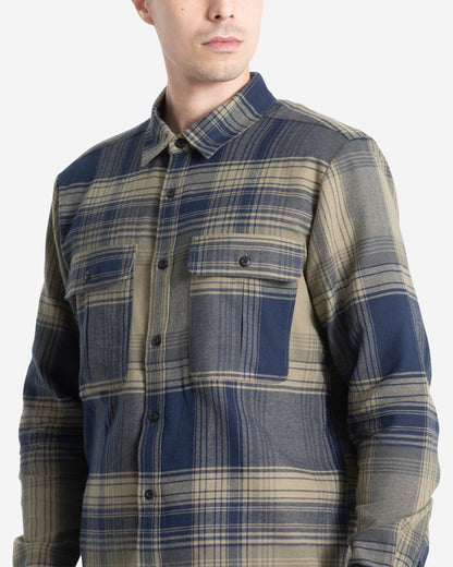A man wears the Volcom Men's Parkstone Long Sleeve Shirt, a blue and beige plaid button-up with two chest pockets, standing against a plain light background.
