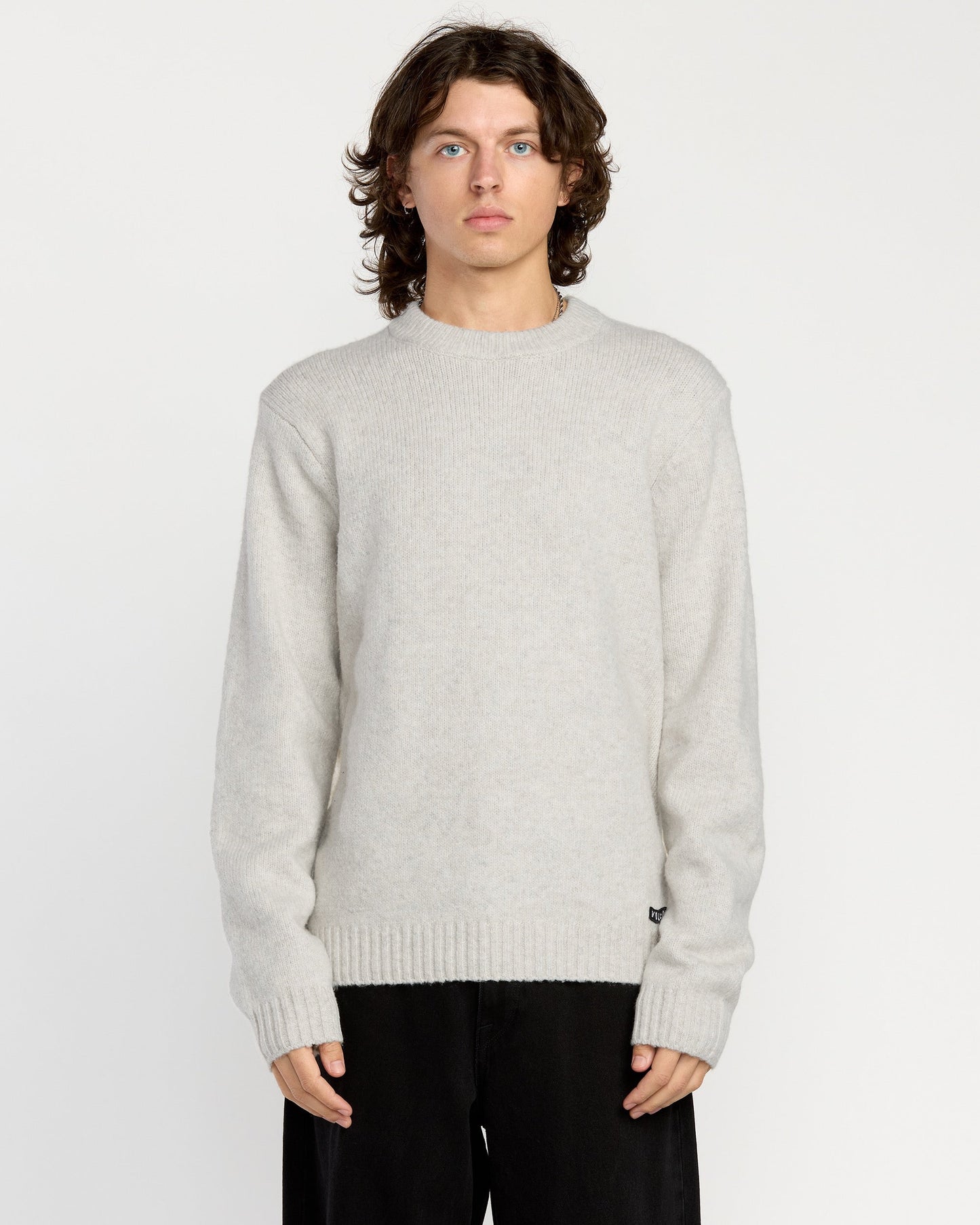 A man with wavy brown hair wears the Volcom Men's Edmonder II Sweater in light gray and loose black pants, standing facing forward against a plain white background.