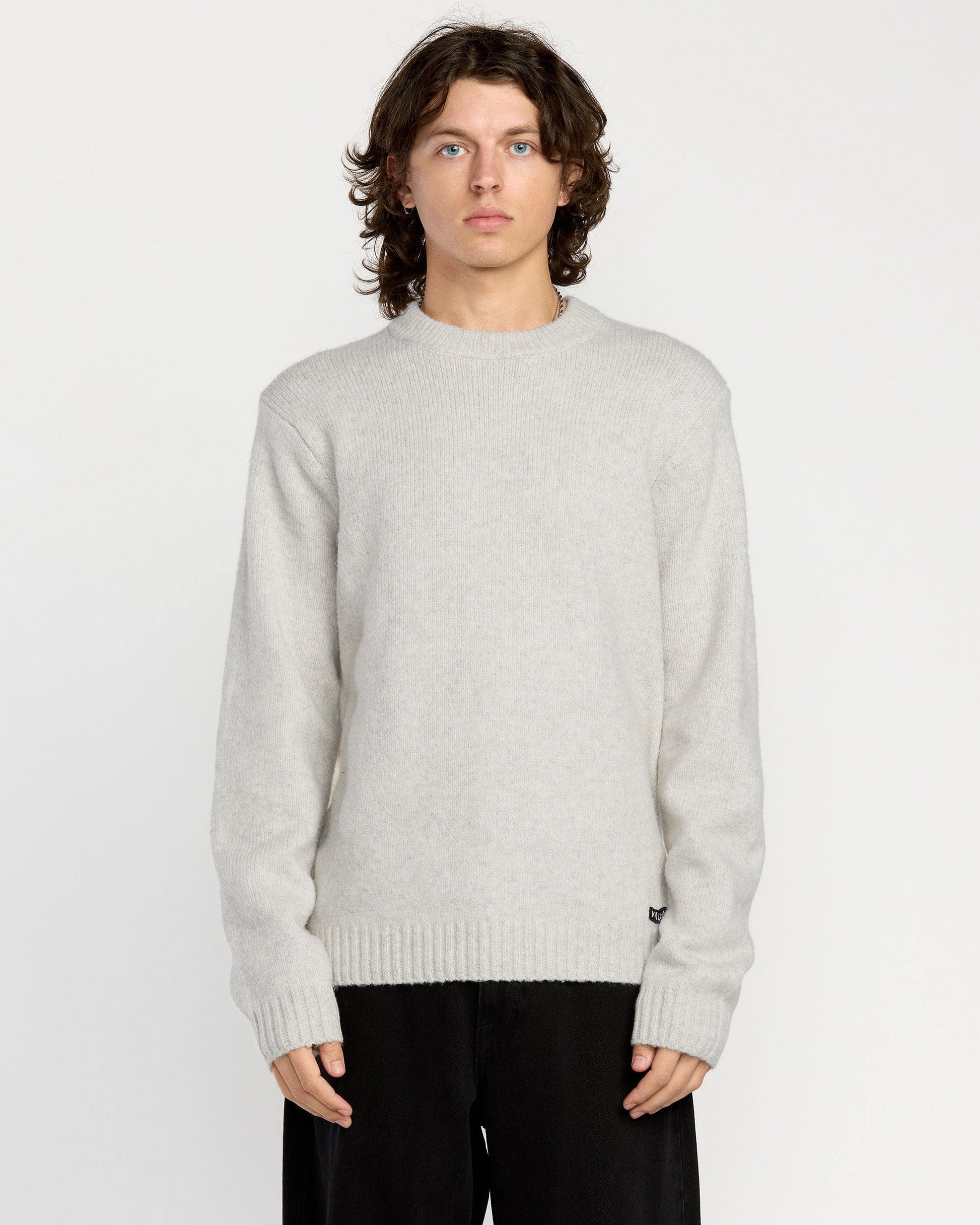 A man with wavy brown hair wears the Volcom Men's Edmonder II Sweater in light gray and loose black pants, standing facing forward against a plain white background.