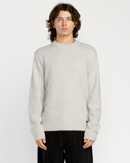 A man with wavy brown hair wears the Volcom Men's Edmonder II Sweater in light gray and loose black pants, standing facing forward against a plain white background.