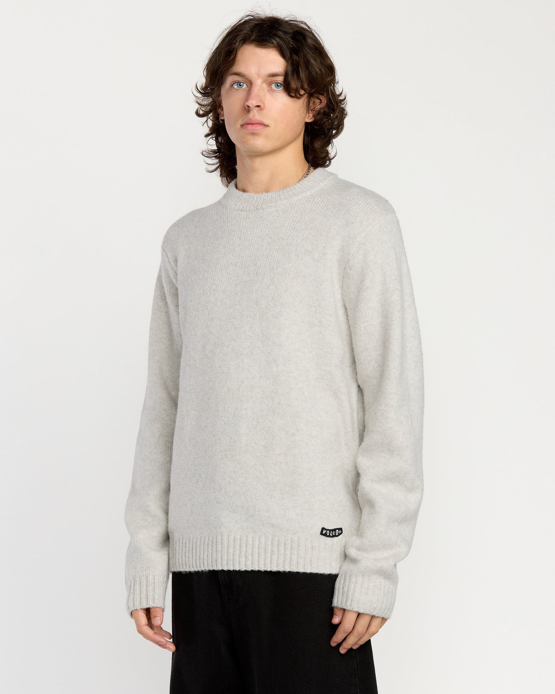 A person with shoulder-length wavy hair wears the Volcom Men's Edmonder II Sweater in light gray and black pants, standing against a plain white background.