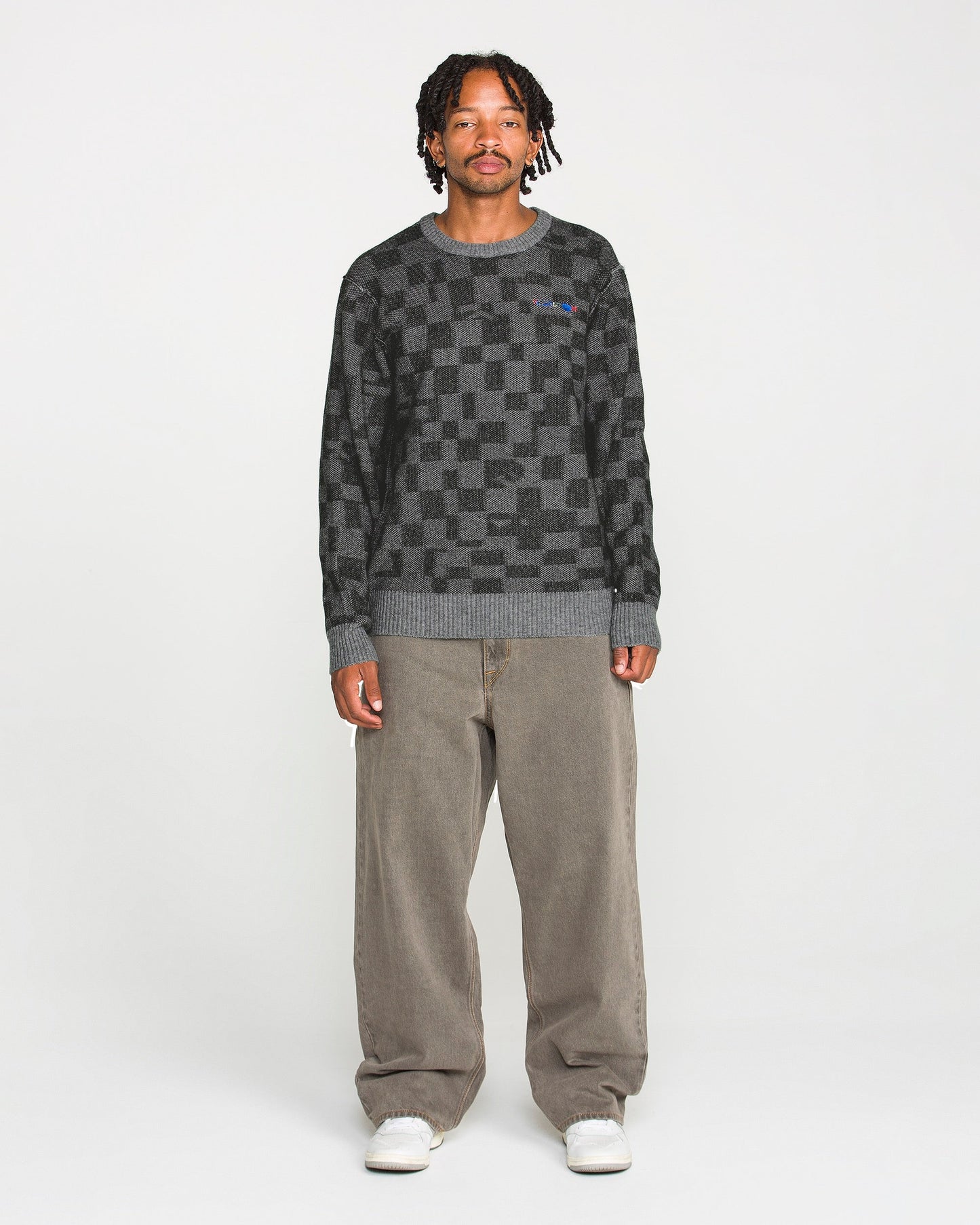 A person stands facing forward against a plain white background, wearing the Volcom Men's Featured Artist Antonio Carrau Sweater with a boxy fit, paired with loose brown pants and white sneakers.