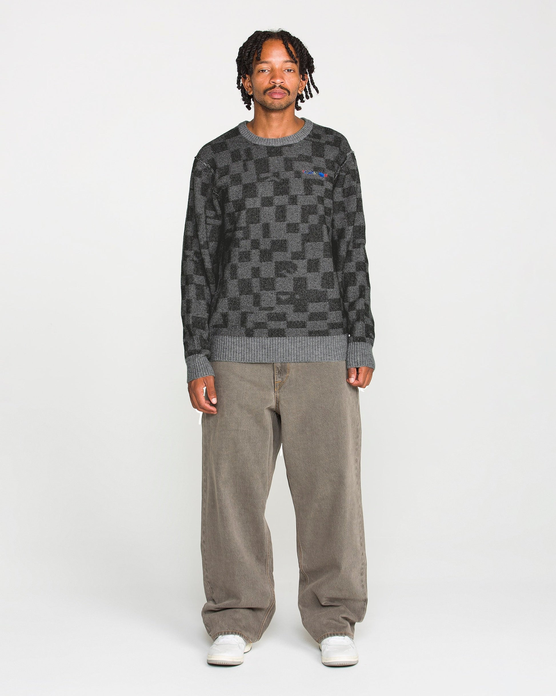 A person stands facing forward against a plain white background, wearing the Volcom Men's Featured Artist Antonio Carrau Sweater with a boxy fit, paired with loose brown pants and white sneakers.
