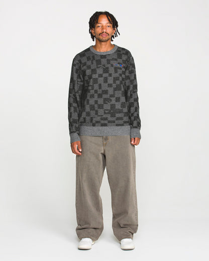 A person stands facing forward against a plain white background, wearing the Volcom Men's Featured Artist Antonio Carrau Sweater with a boxy fit, paired with loose brown pants and white sneakers.