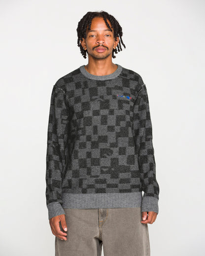 A person with short dreadlocks wears the Volcom Men's Featured Artist Antonio Carrau Sweater—black and gray jacquard—in addition to loose gray pants, standing against a plain white background and facing the camera with a neutral expression.