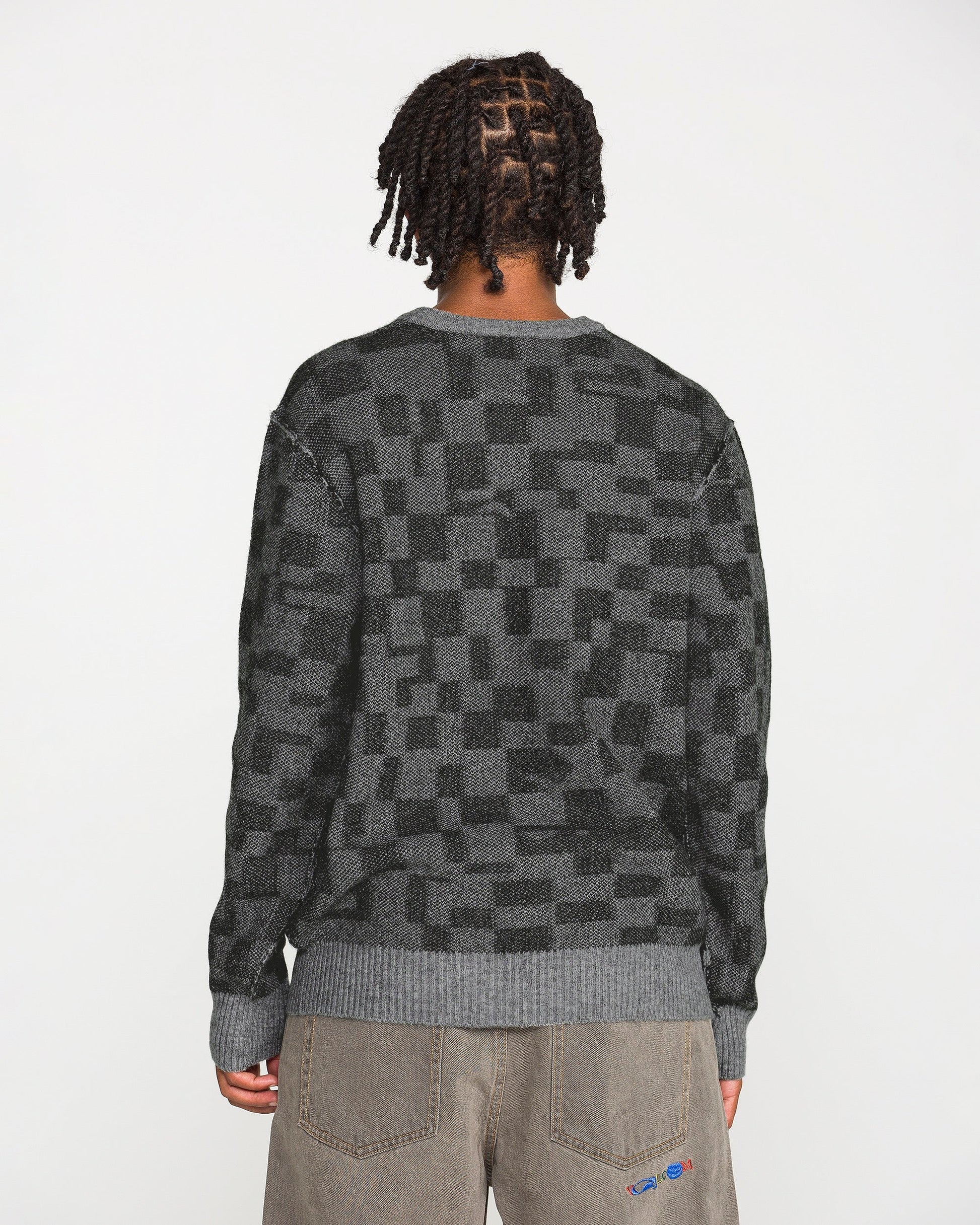 A person with short, twisted hair is seen from behind against a plain white background, wearing the Volcom Men's Featured Artist Antonio Carrau Sweater in dark gray checkered jacquard paired with brown pants.