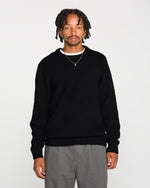 A man with short dreadlocks wears the Volcom Men's Edmonder II Sweater in black, paired with gray pants and a gold necklace. He stands facing forward against a plain white background.
