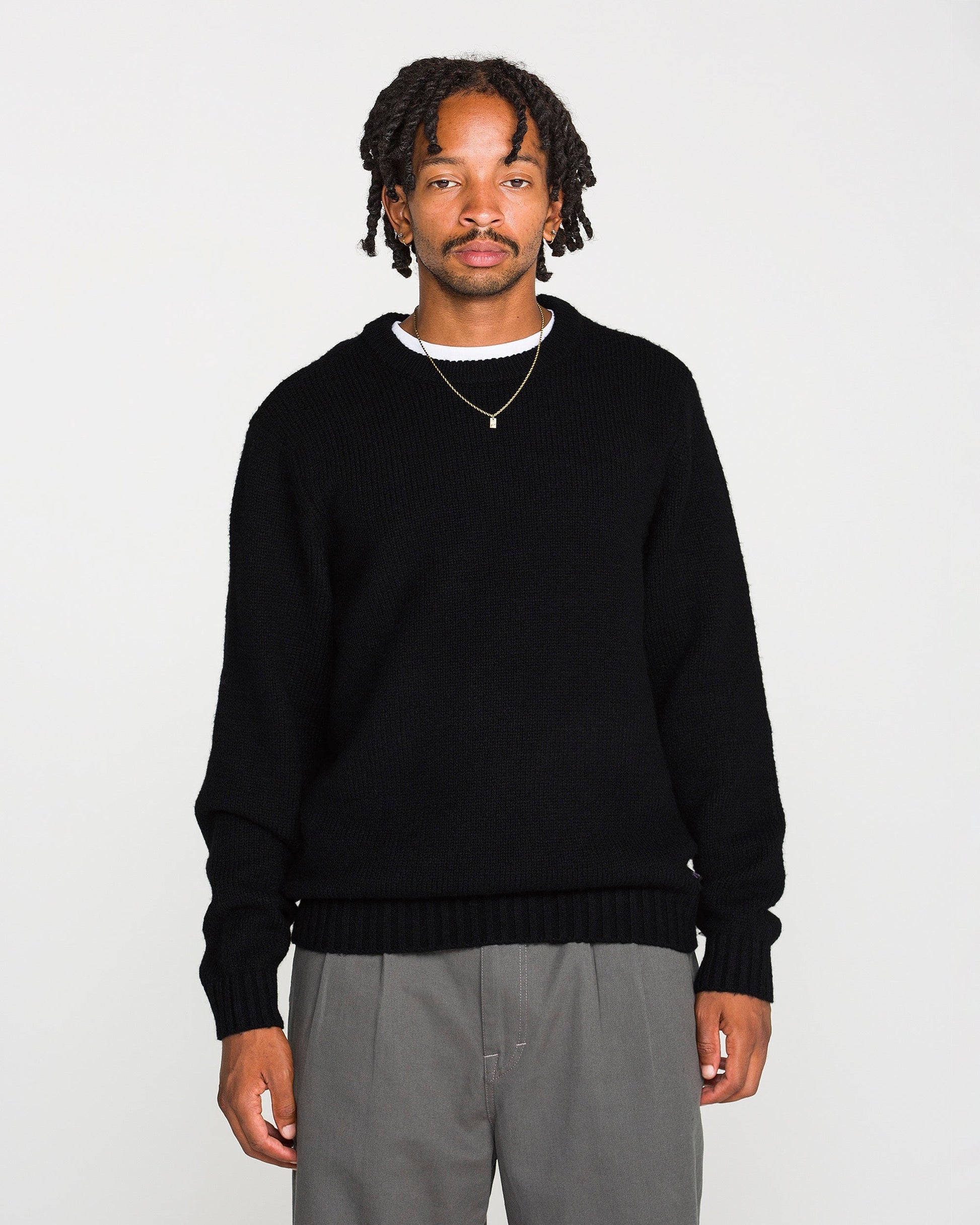 A man with short dreadlocks wears the Volcom Men's Edmonder II Sweater in black, paired with gray pants and a gold necklace. He stands facing forward against a plain white background.