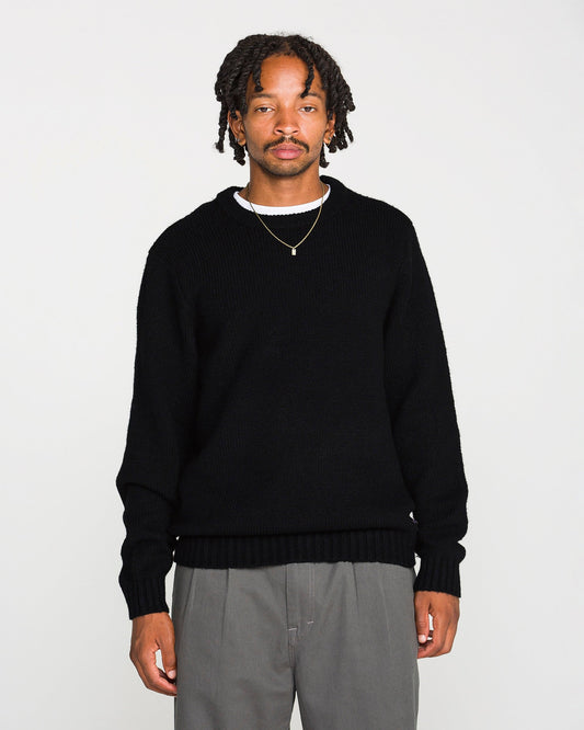 A man with short dreadlocks wears the Volcom Men's Edmonder II Sweater in black, paired with gray pants and a gold necklace. He stands facing forward against a plain white background.