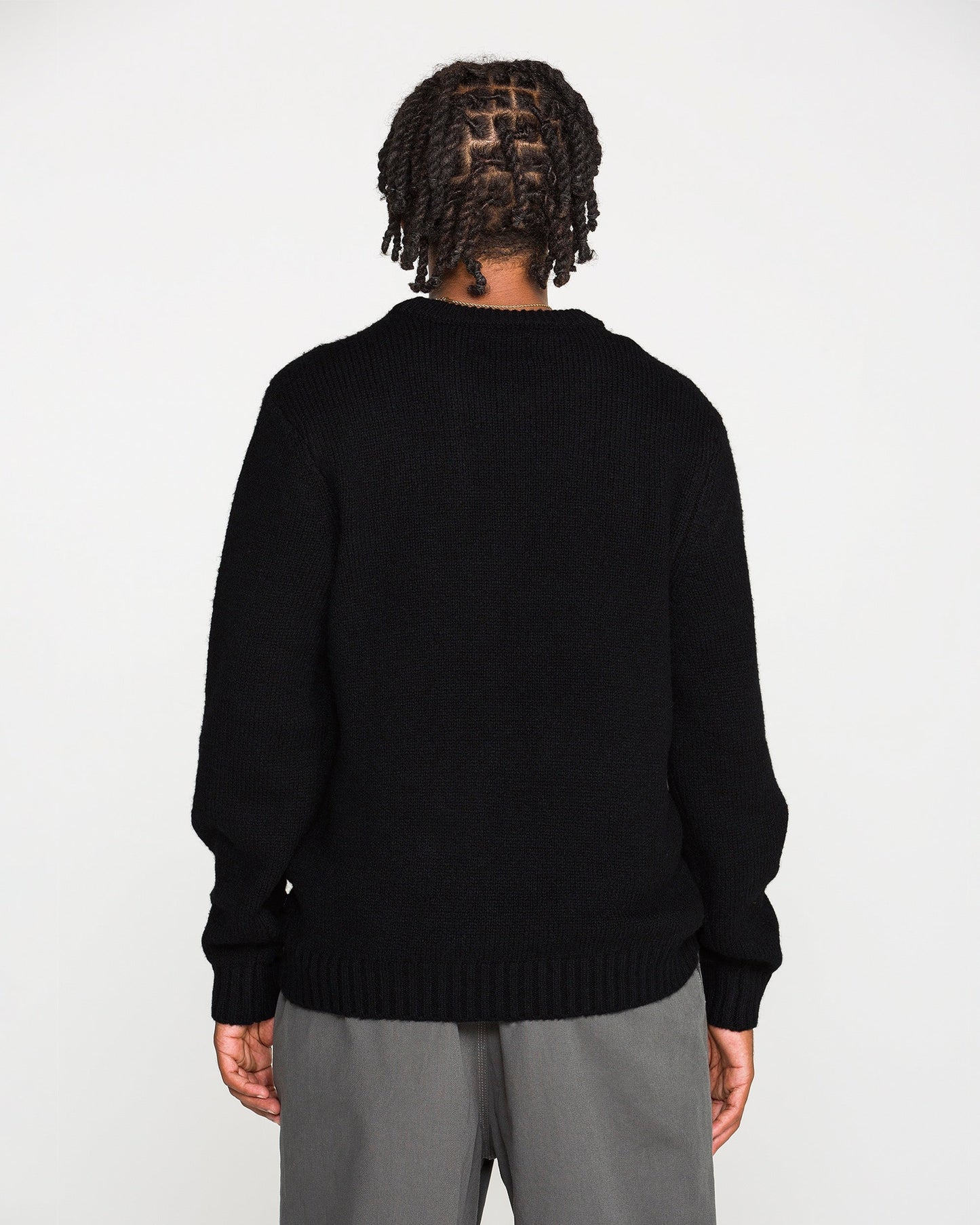A person with braided hair stands facing away, wearing the Volcom Men's Edmonder II Sweater in black—crafted from recycled polyester—paired with gray pants, against a plain white background.