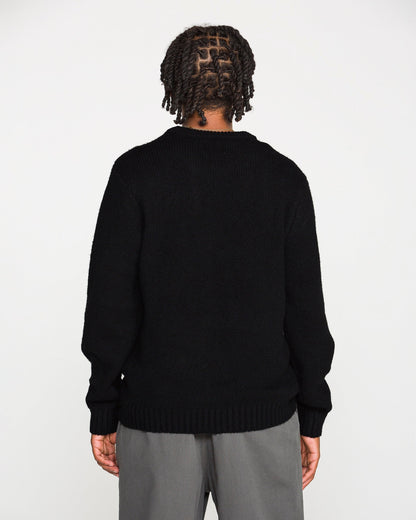 A person with braided hair stands facing away, wearing the Volcom Men's Edmonder II Sweater in black—crafted from recycled polyester—paired with gray pants, against a plain white background.