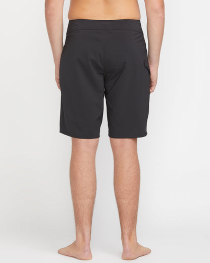 A person stands barefoot, facing away from the camera, wearing Volcom Men's Lido Solid Mod 20 black knee-length shorts against a plain white background.