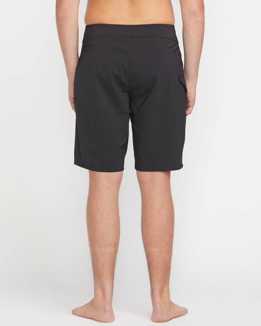 A person stands barefoot, facing away from the camera, wearing Volcom Men's Lido Solid Mod 20 black knee-length shorts against a plain white background.