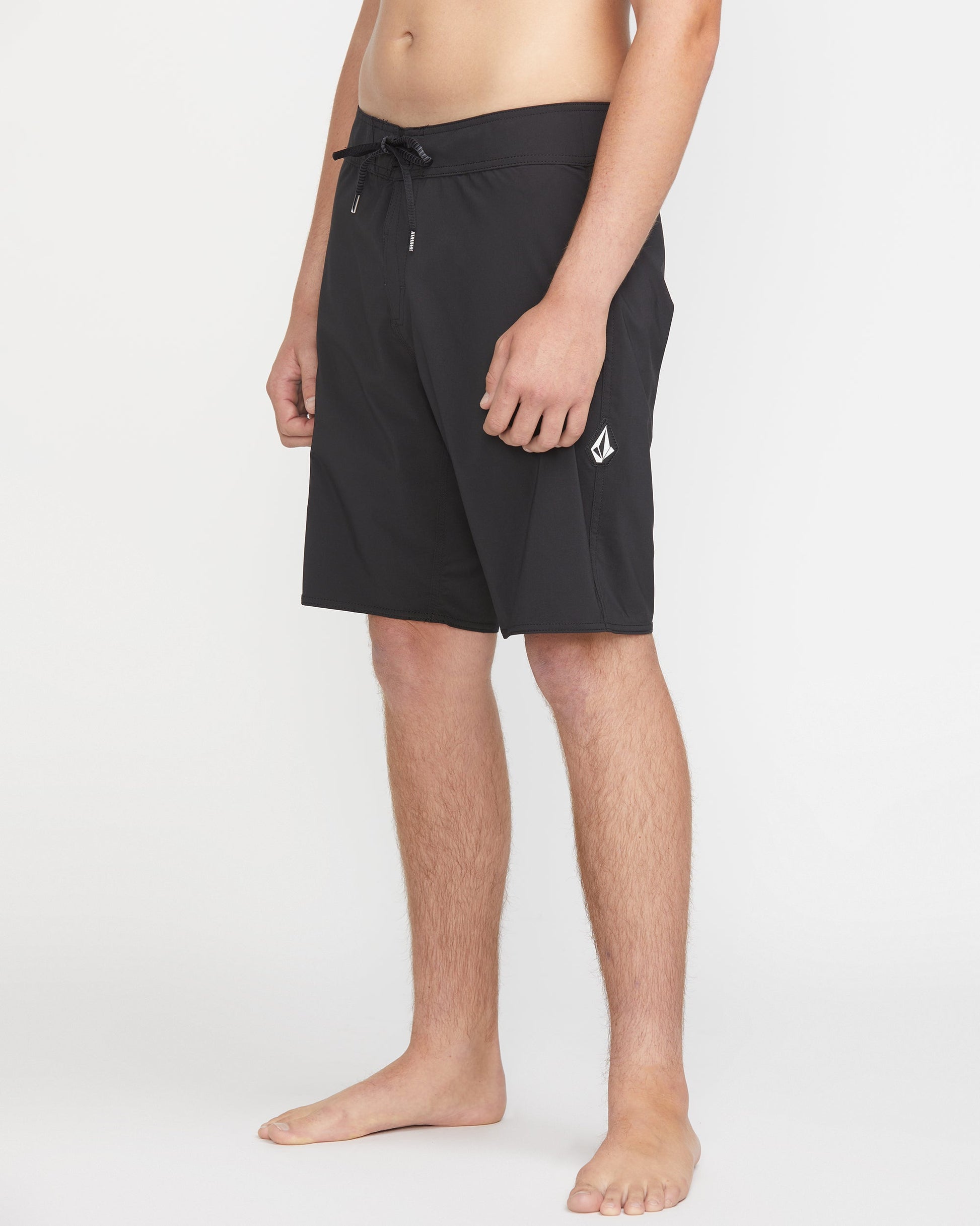 A man wears Volcom Men's Lido Solid Mod 20 black knee-length swim trunks with a drawstring waist and a small white logo on the left leg, standing barefoot against a plain white background.