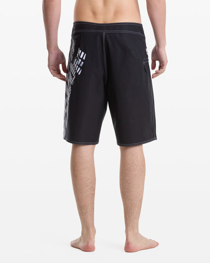 A man stands barefoot, facing away from the camera, wearing Volcom Men's Reaper Mod 21 knee-length black shorts with white stitching and a geometric white pattern on the left side against a plain white background.