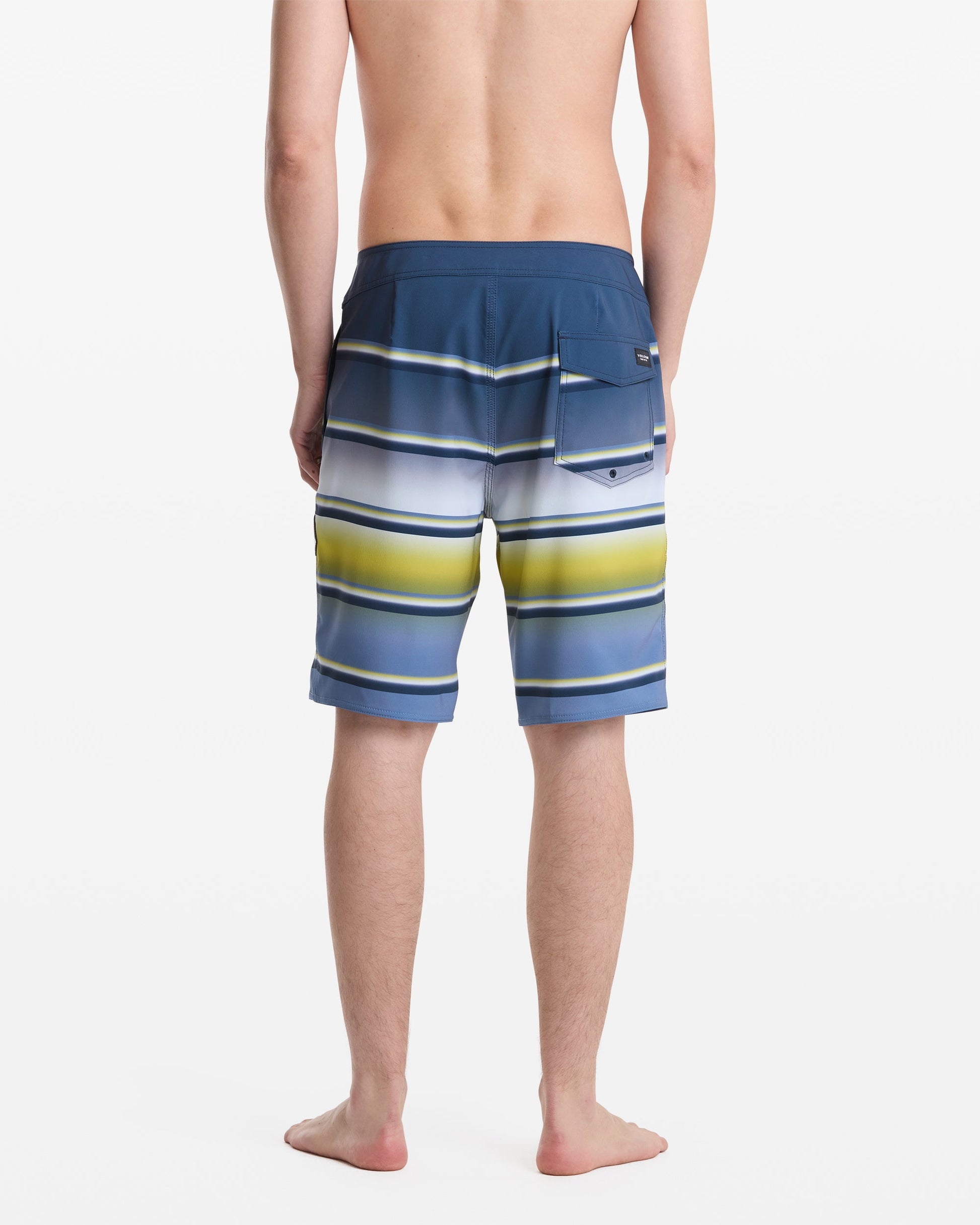 A man stands barefoot with his back to the camera, wearing Volcom Men's Quarta Dissolve Mod 20 blue board shorts featuring horizontal white, yellow, and green stripes and a right-side pocket.