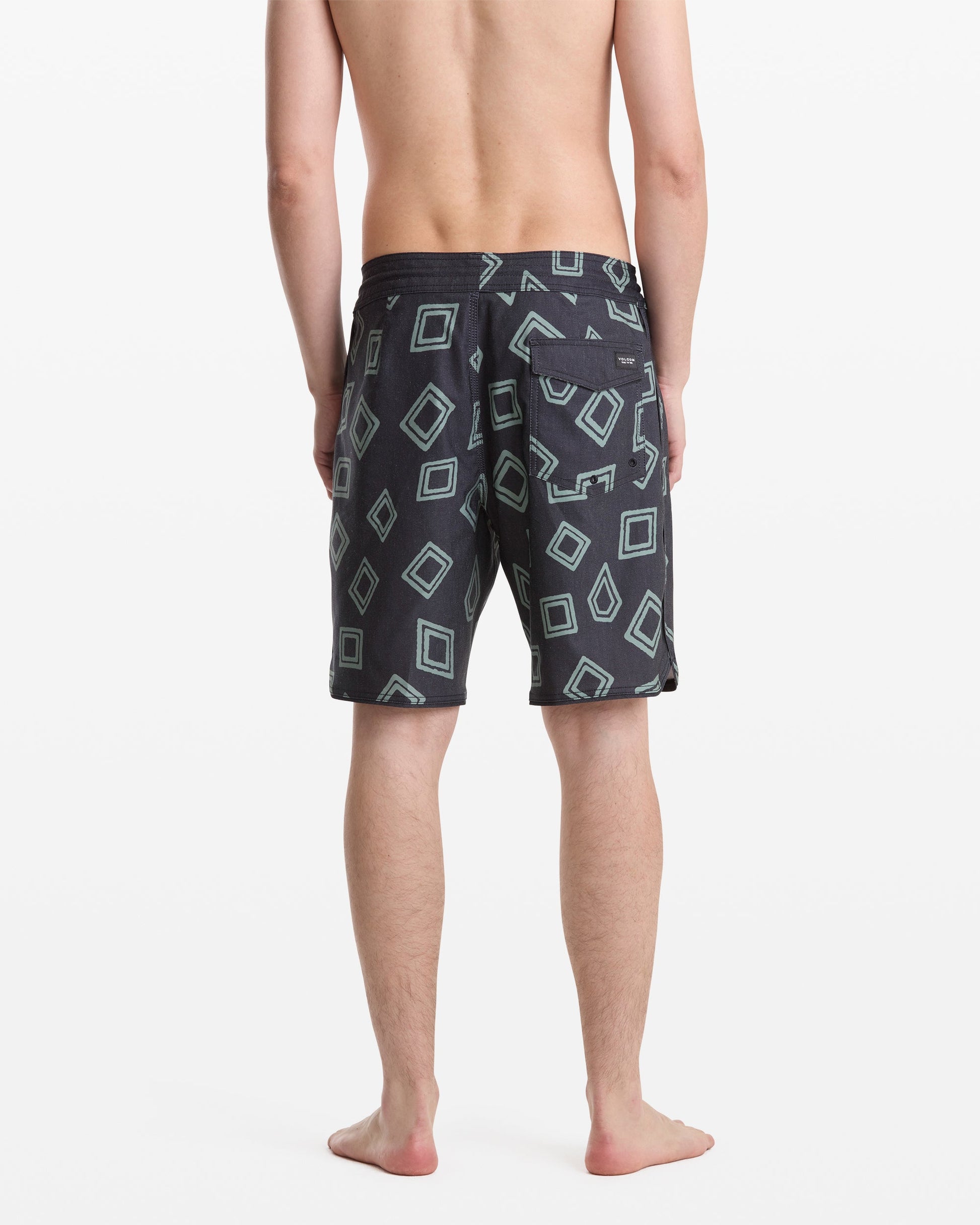A man stands barefoot facing away, wearing Volcom Men's Contact Scallop Stoney 19 swim trunks in dark color with light blue geometric square patterns on a white background.
