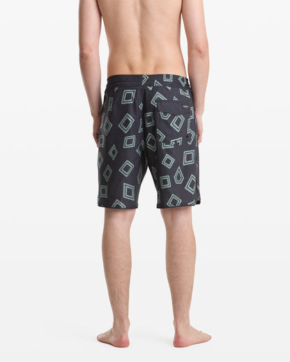 A man stands barefoot facing away, wearing Volcom Men's Contact Scallop Stoney 19 swim trunks in dark color with light blue geometric square patterns on a white background.