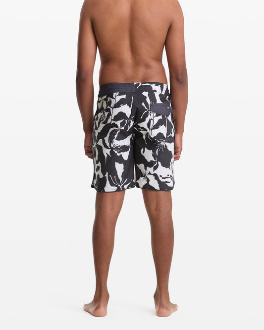 A man stands barefoot facing away, wearing Volcom Men's Contact Scallop Stoney 19 swim trunks with a black and white floral pattern, set against a plain white background.