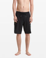 A barefoot man stands against a white backdrop, wearing Volcom Men's Hawaii Top Tier Mod black board shorts with side designs and a drawstring waist. His upper body is unclothed.