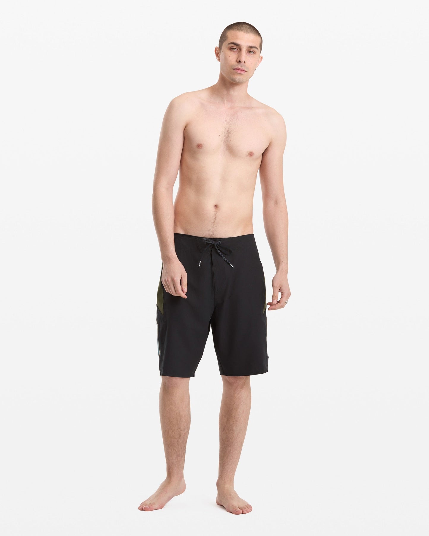 A young man with short hair stands barefoot against a white background, wearing Volcom Men's Hawaii Top Tier Mod black knee-length board shorts with a drawstring and no shirt.