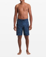A man stands barefoot against a white background, wearing Volcom Men's Shoreline Mod 20 dark blue patterned board shorts with no shirt. His arms are relaxed at his sides as he faces forward.