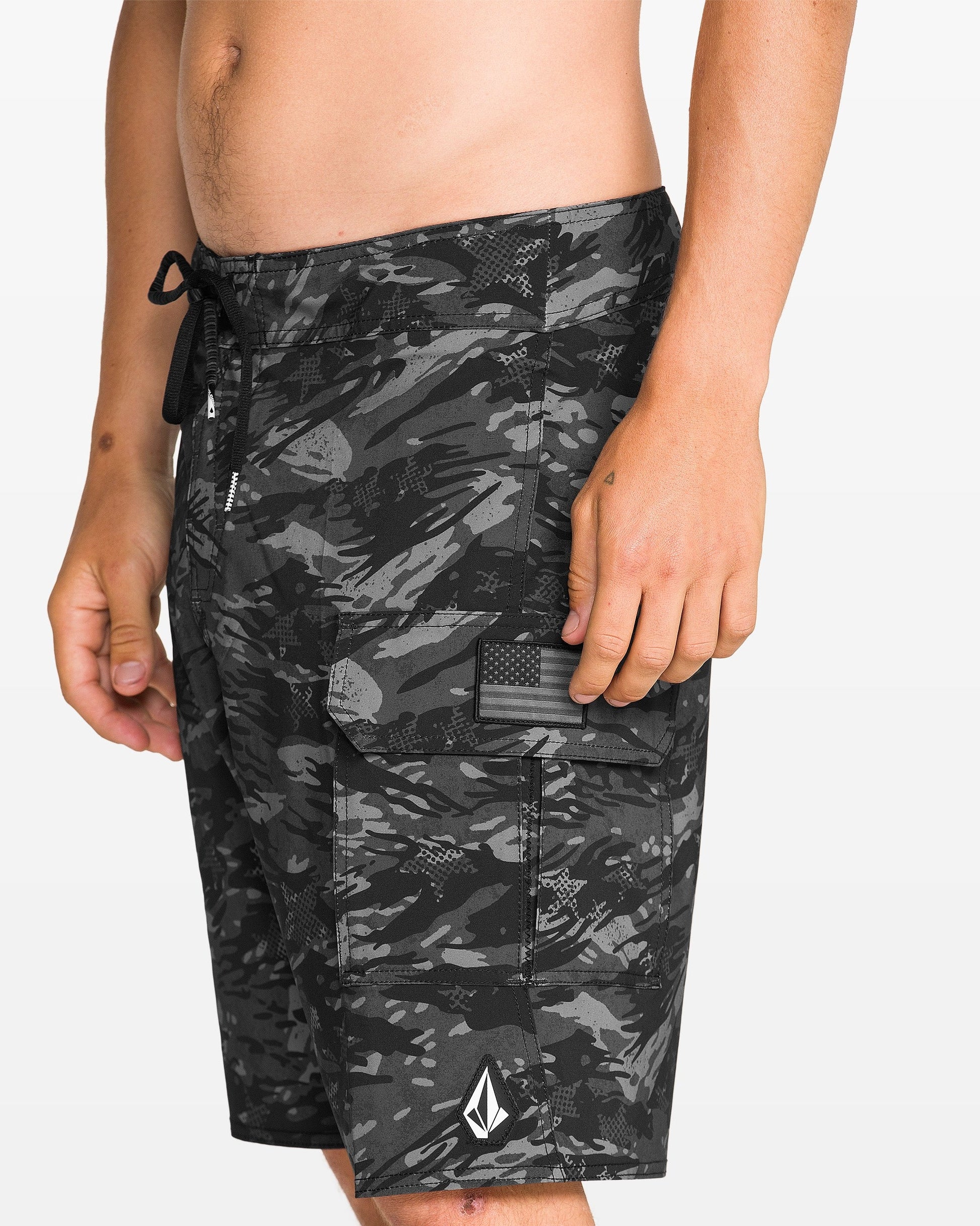 A person wearing Volcom Men's 4th Of July Preemo Mod black and gray camo boardshorts with Cinch Fly Technology, a side cargo pocket with Velcro flap, a monochrome American flag patch, and a white logo near the hem.