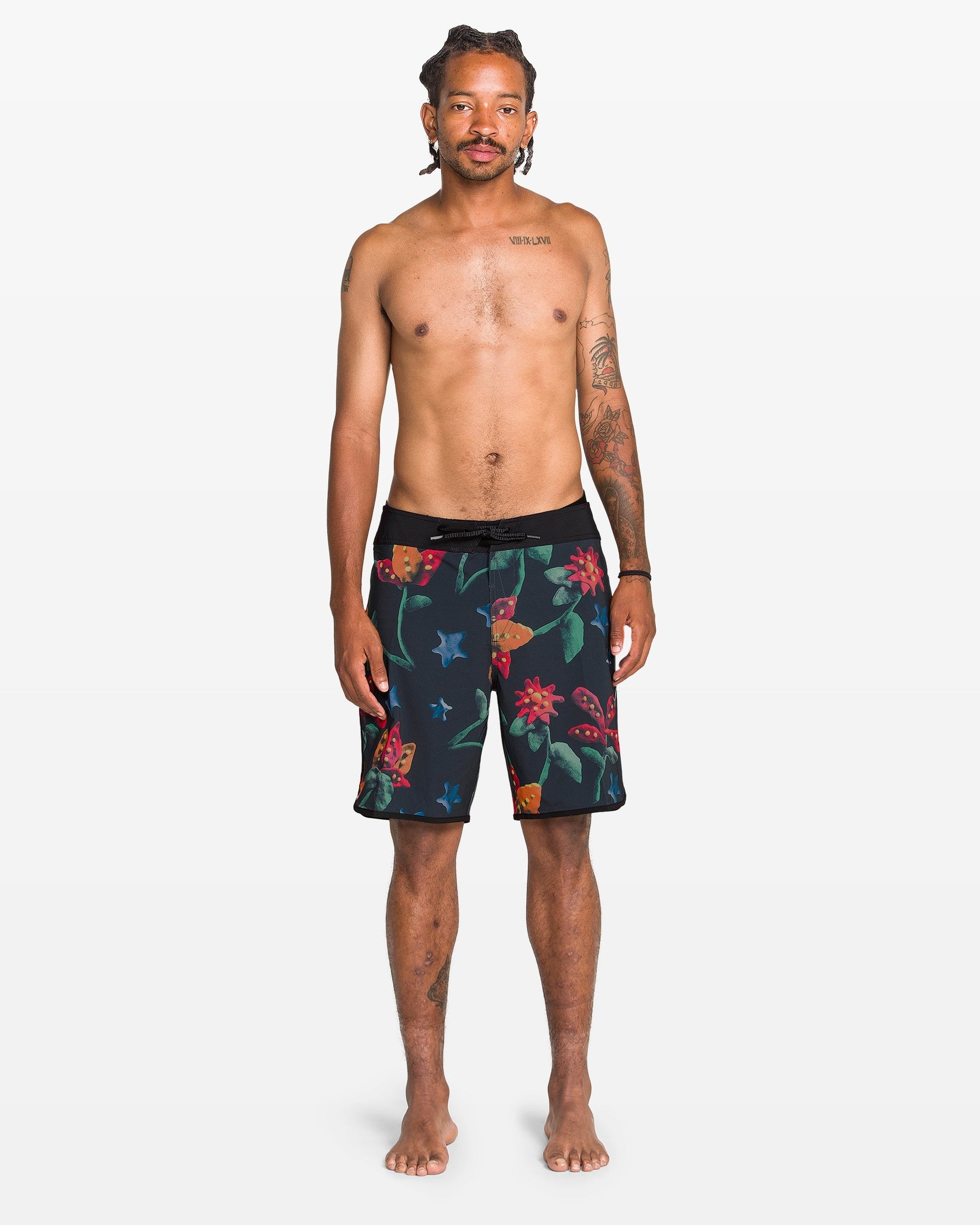 A tattooed man stands barefoot against a white background, facing forward in Volcom Men's Infuse Scallop Mod Boardshorts, which feature a colorful floral pattern and Cinch Fly Technology. He is shirtless with a neutral expression.