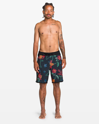 A tattooed man stands barefoot against a white background, facing forward in Volcom Men's Infuse Scallop Mod Boardshorts, which feature a colorful floral pattern and Cinch Fly Technology. He is shirtless with a neutral expression.