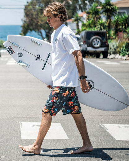 A young man wearing Volcom's Men's Infuse Scallop Mod boardshorts and a white t-shirt walks barefoot across a street with a surfboard, while palm trees, parked cars, and the ocean appear in the background.