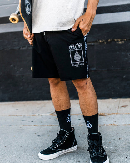 A person wearing Volcom Men's Yewster Loose Stoney black boardshorts, black socks, and black sneakers stands on a sidewalk holding a skateboard. Only the lower half of their body is visible.