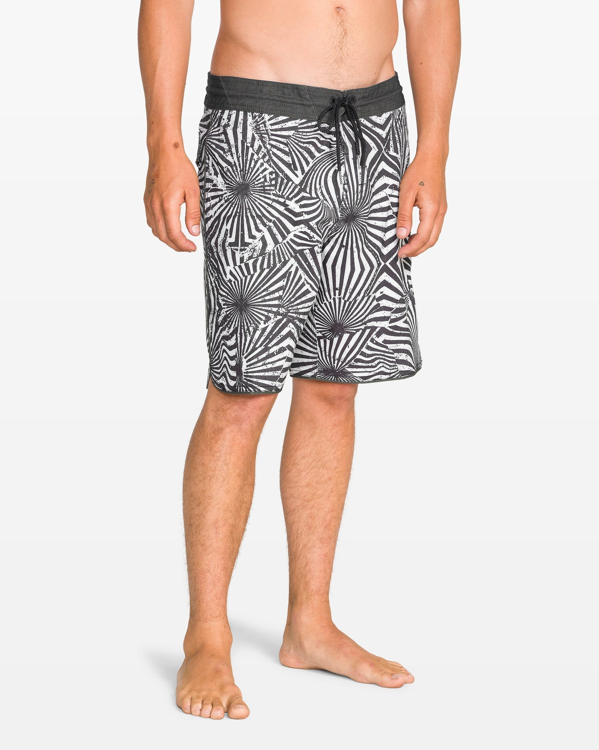 A man stands barefoot against a plain white background, wearing Volcom Men's Barnacle Scallop Stoney black and white patterned boardshorts featuring the Stoney Cinch Fly.