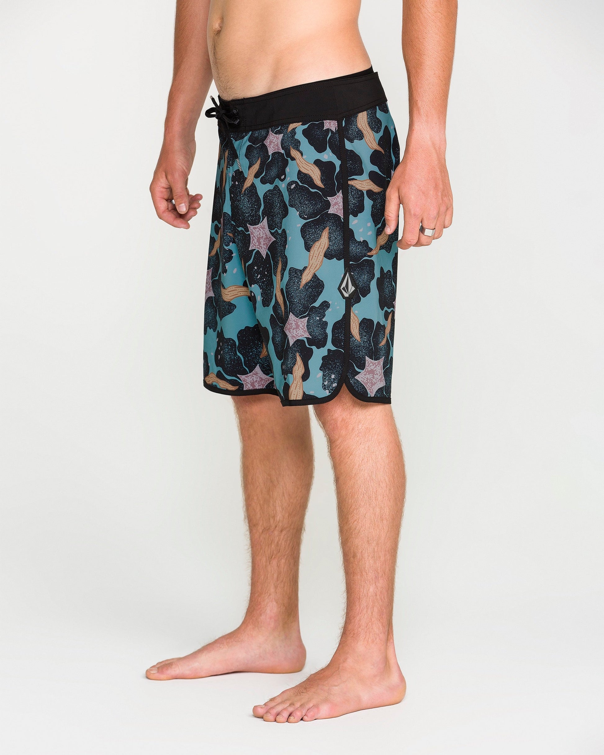 A person stands barefoot on a white background, wearing Volcom Men's Infuse Scallop Mod board shorts featuring a teal and black pattern, drawstring waist, and 4-way stretch for maximum mobility. Only the lower half of the body is visible.