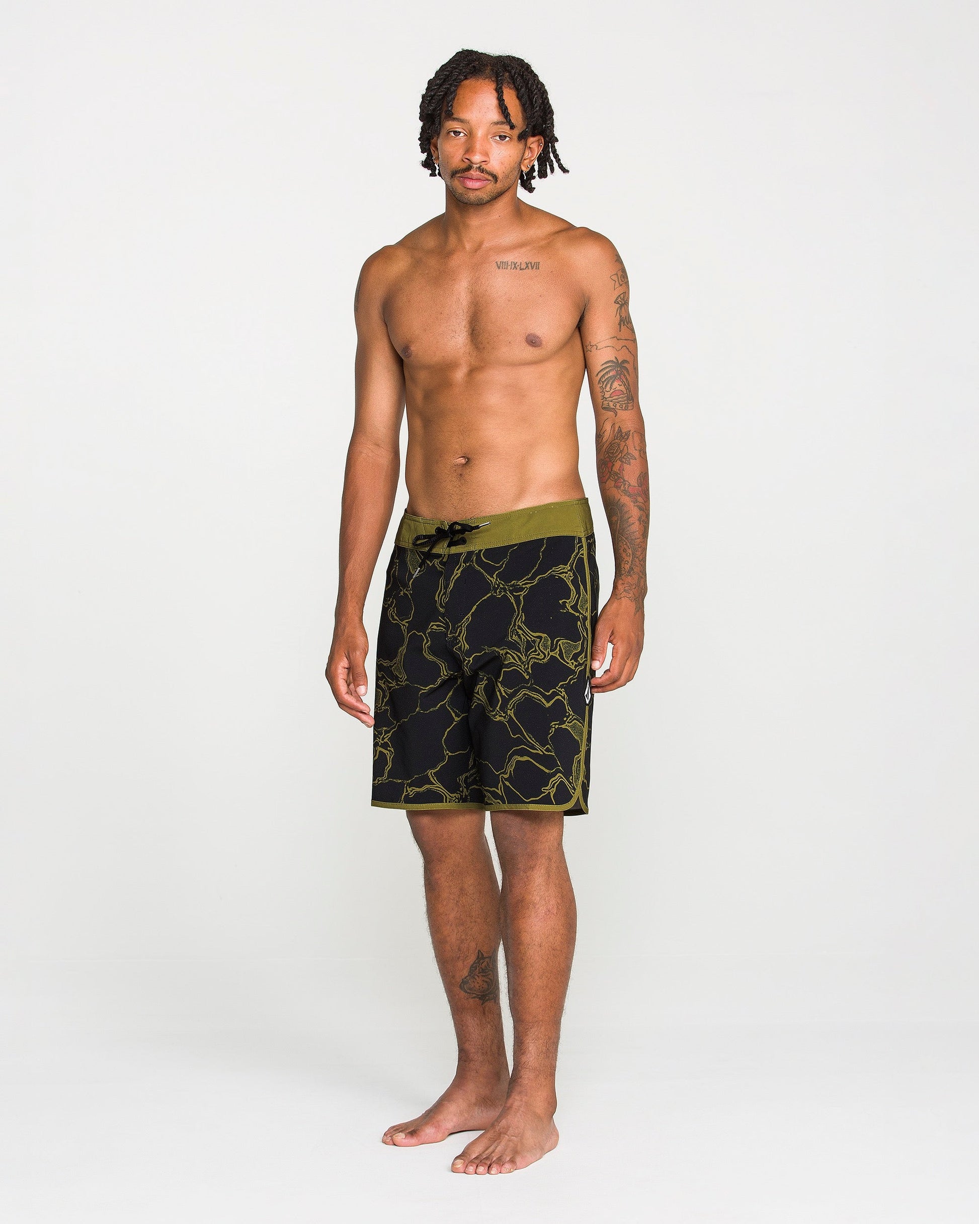 A man with short dreadlocks and tattoos stands barefoot, wearing Volcom Men's Infuse Scallop Mod boardshorts in black and olive green floral 4-way stretch fabric with a Cinch Fly, against a plain white background.