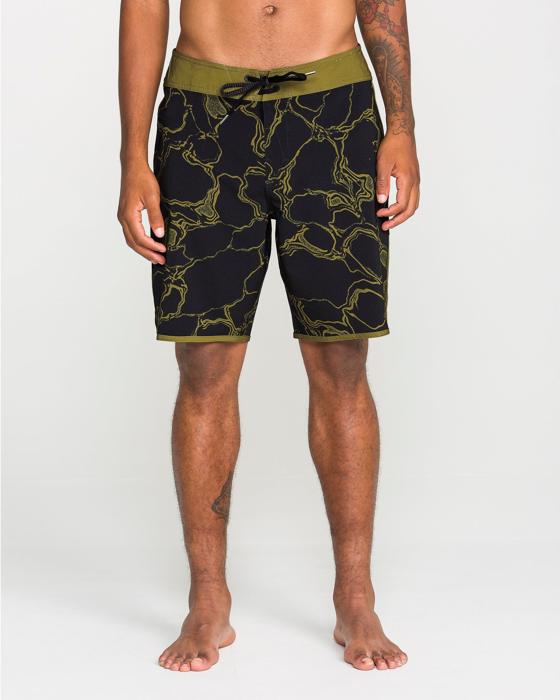 A person stands barefoot against a plain white background, hands relaxed at their sides, wearing the Volcom Men's Infuse Scallop Mod boardshorts in black and olive-green with a yellow marble pattern and 4-way stretch for enhanced comfort.