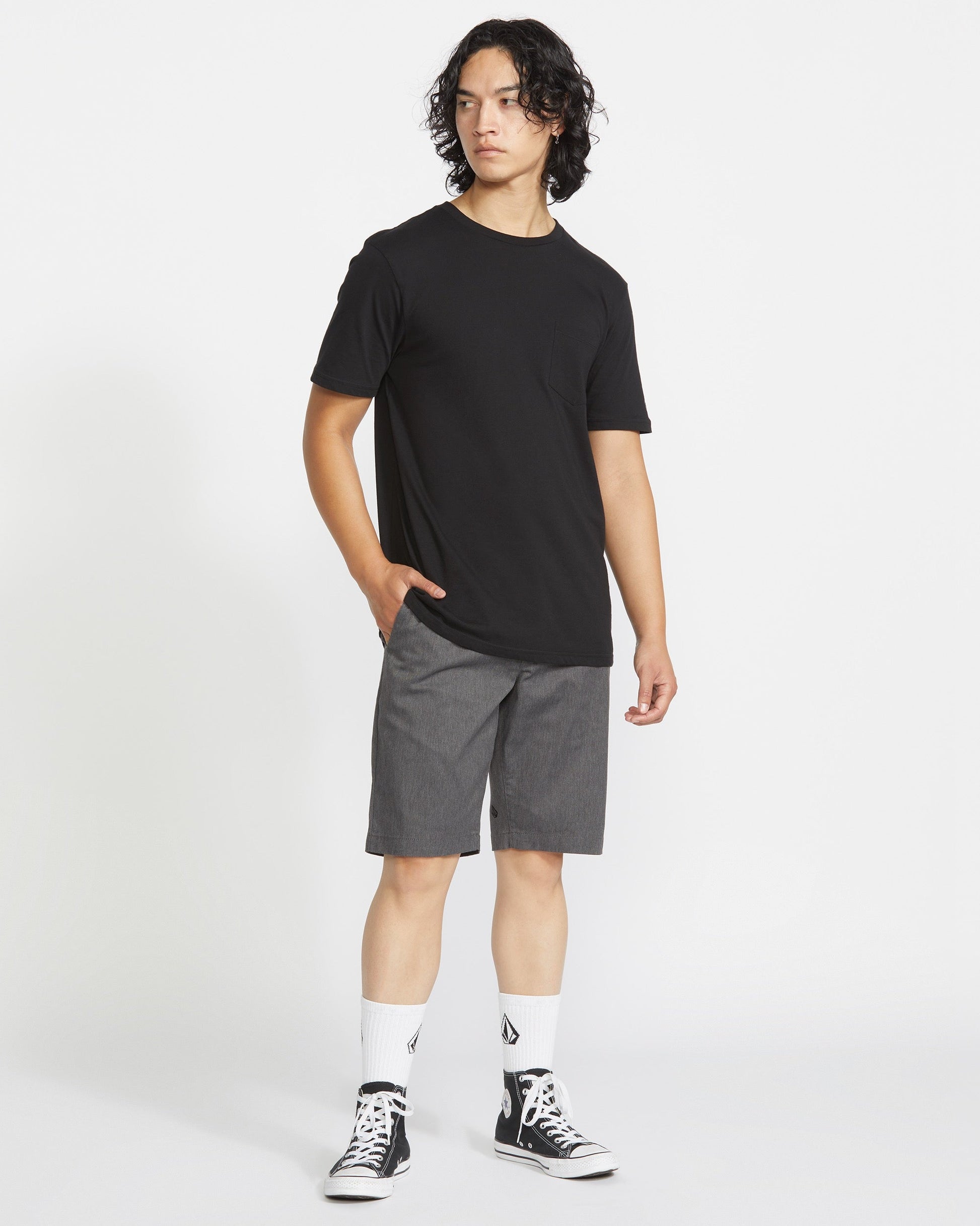 A person stands against a plain white background wearing a Volcom black t-shirt, Men's Frickin Modern Stretch Shorts by Volcom in breathable gray cotton, white socks with black logos, and black high-top sneakers. One hand rests in their pocket as they look sideways.